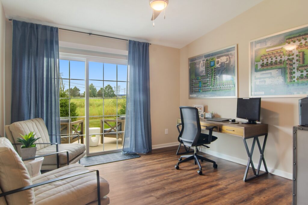 Home office with a desk, computer, and chair beside two armchairs; large window with blue curtains overlooks a grassy outdoor area.