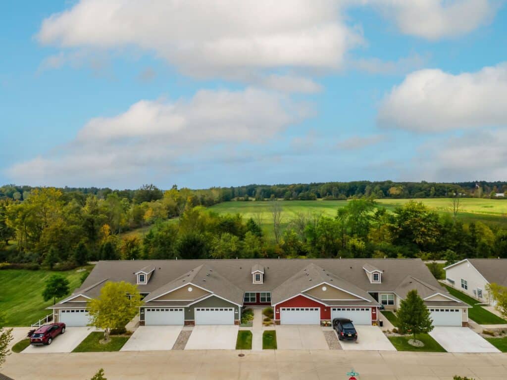 A row of attached suburban houses with garages faces a street, with green fields and trees in the background under a partly cloudy sky.
