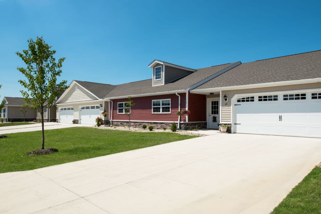 Row of modern single-family homes with attached garages, red and beige siding, manicured lawns, and clear blue sky.