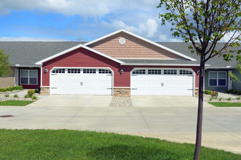 A duplex building with red siding, stone accents, and two white double garages, set against a clear sky with a driveway and a young tree in the foreground.