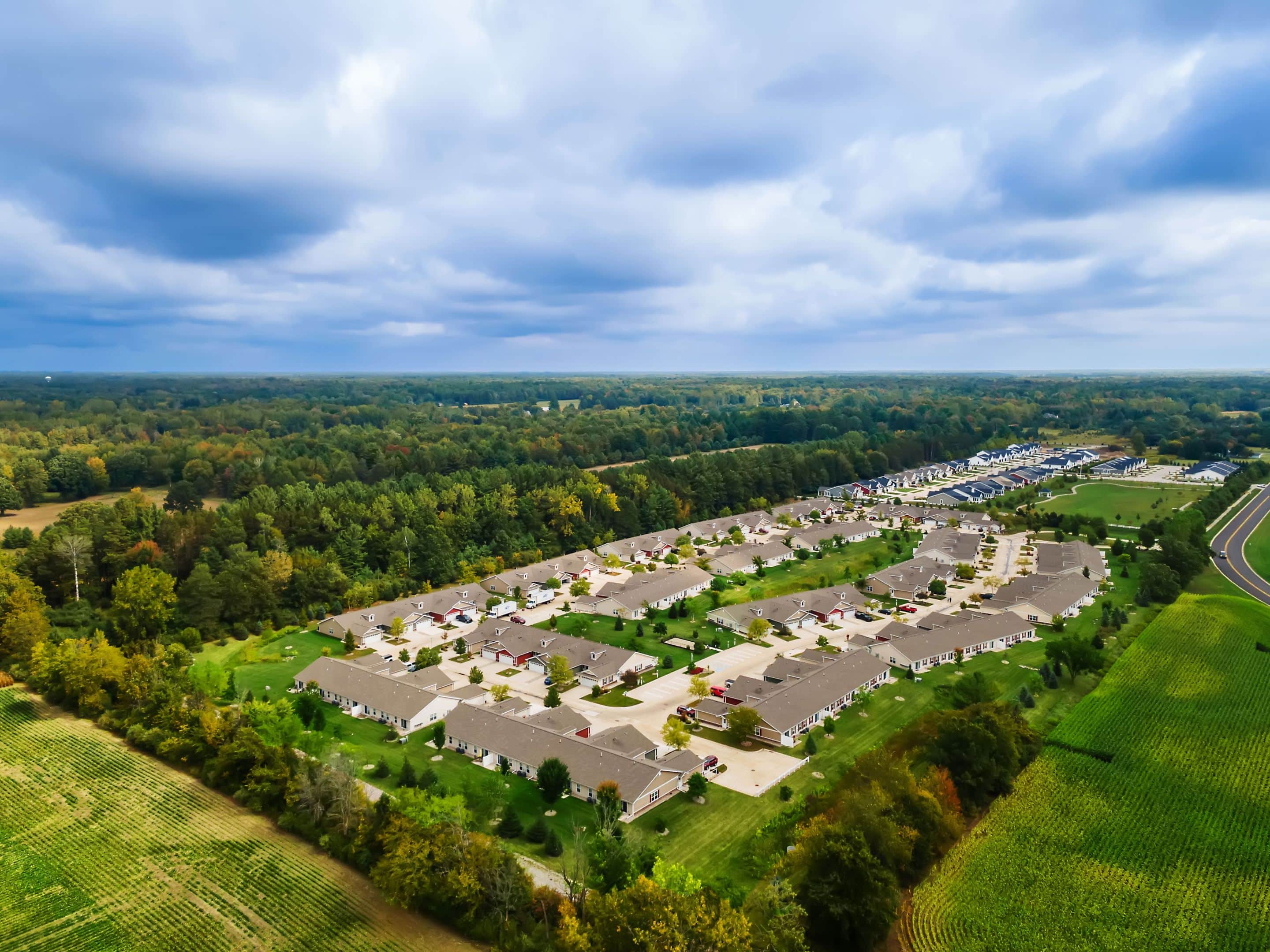 Aerial view of a residential housing development surrounded by green fields and dense forest under a cloudy sky.