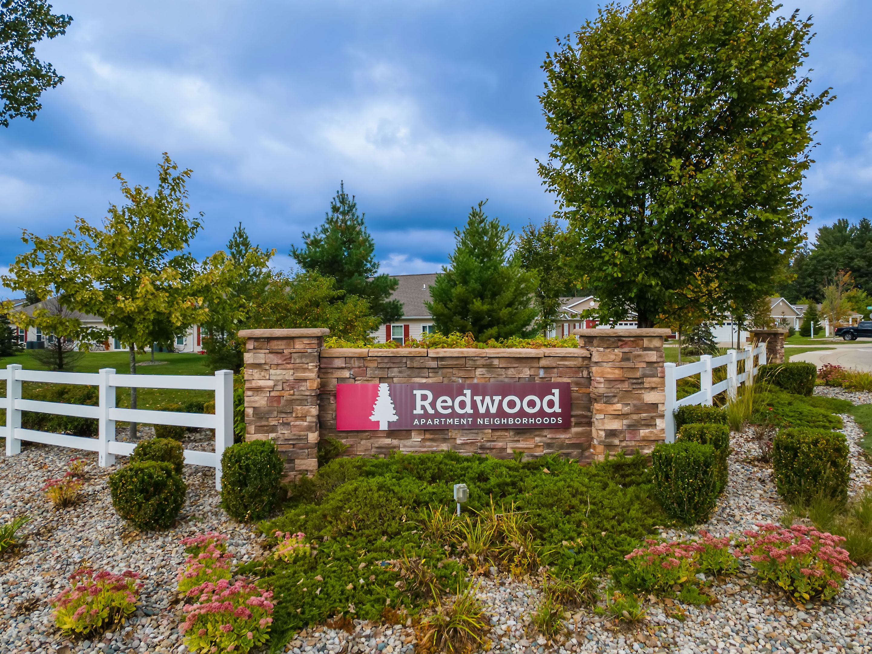 A stone sign reads "Redwood Apartment Neighborhoods" surrounded by landscaping, white fence, trees, and residential buildings in the background under a cloudy sky.