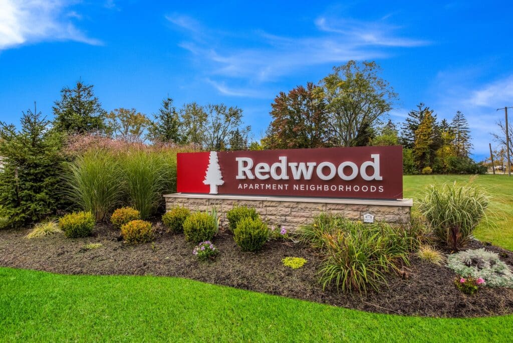 A landscaped area with a large sign reading "Redwood Apartment Neighborhoods" set against green grass, trees, and a blue sky.