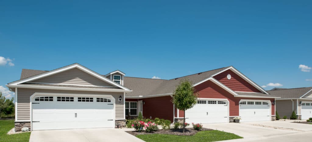Row of single-story suburban houses with attached garages, light and red siding, manicured lawns, and a clear blue sky.