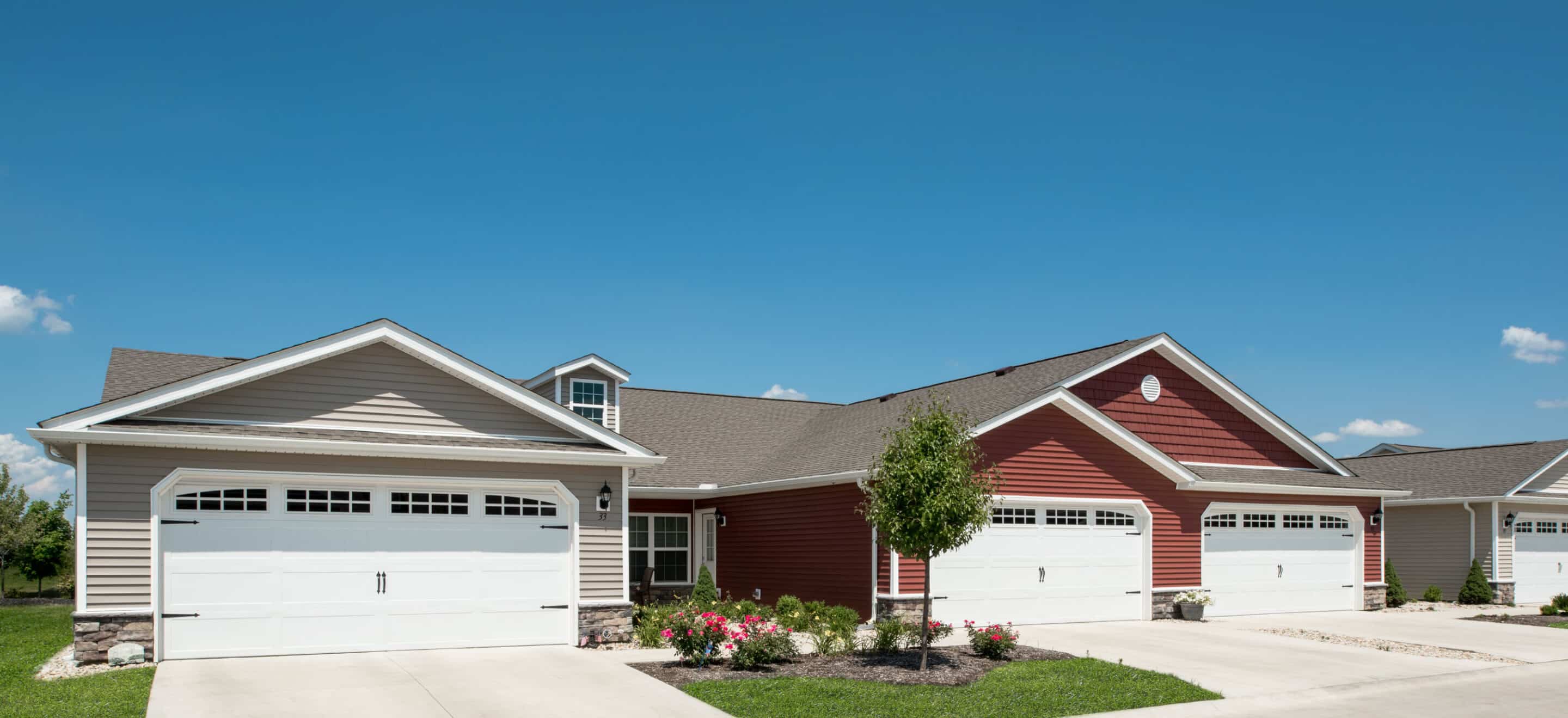 Row of single-story suburban houses with attached garages, light and red siding, manicured lawns, and a clear blue sky.