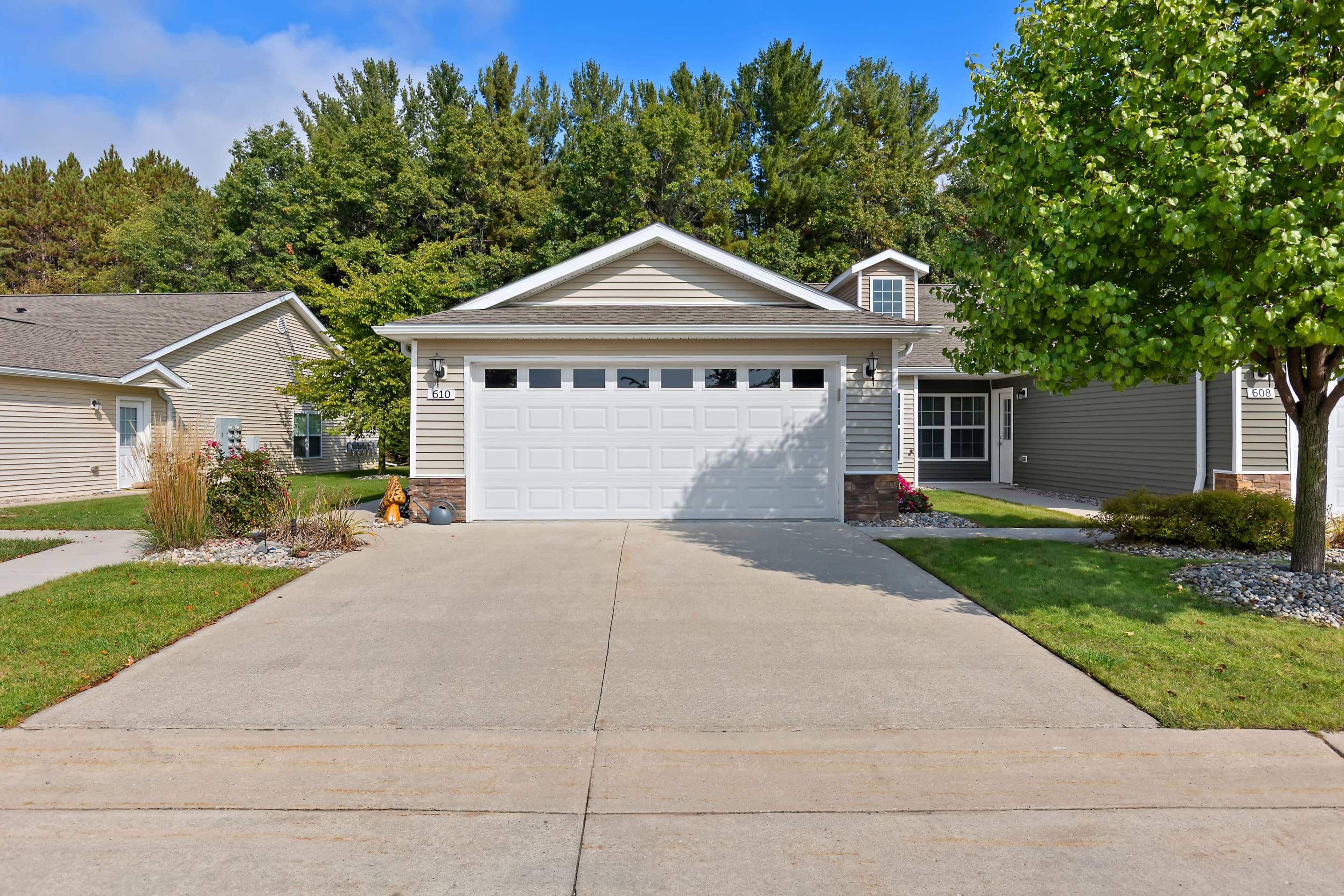 Single-story suburban house with a two-car garage, a driveway, and a well-kept lawn, set against a backdrop of trees and neighboring homes.
