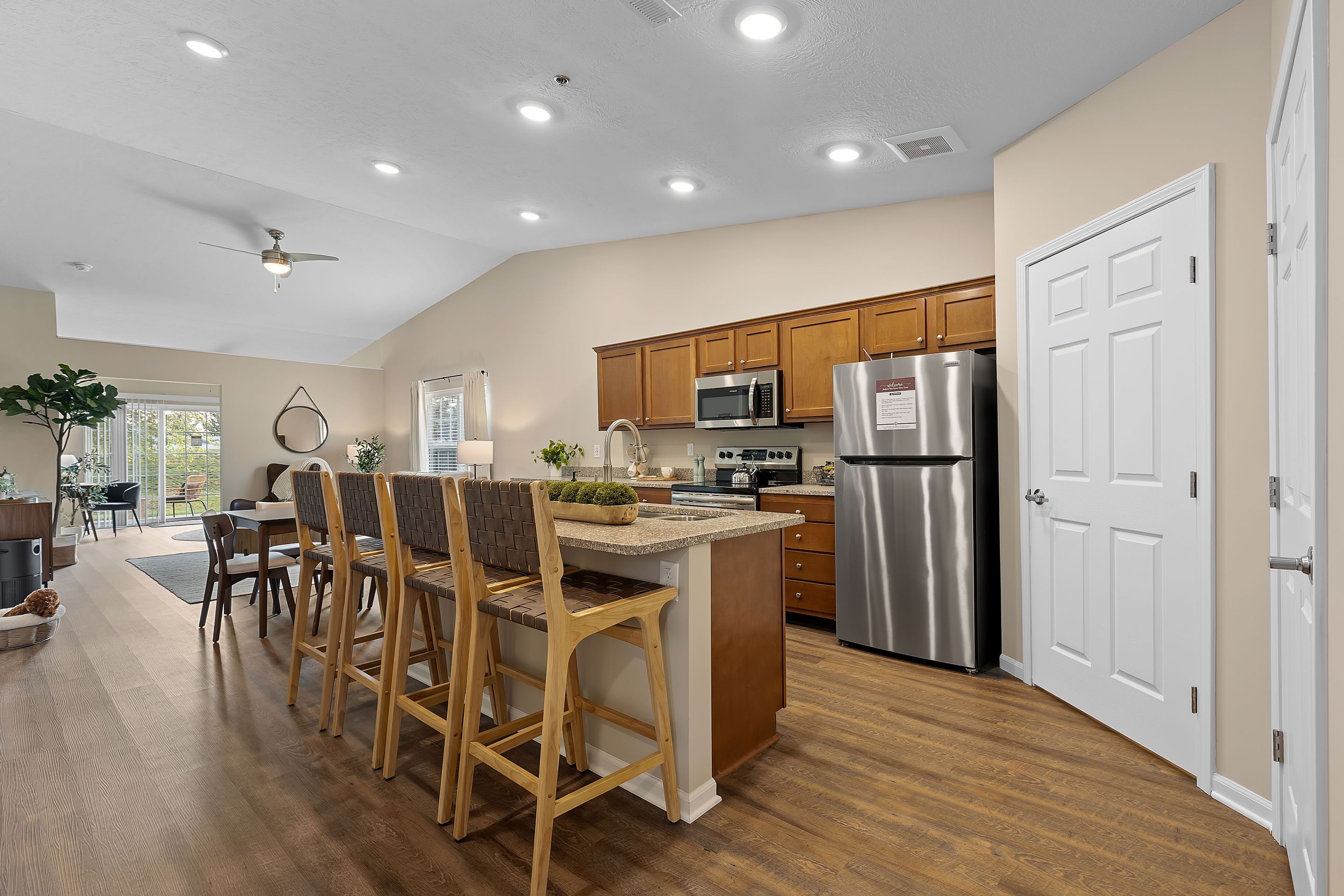 Modern kitchen with wood floors, island seating, stainless steel appliances, brown cabinets, and dining area visible in the background.