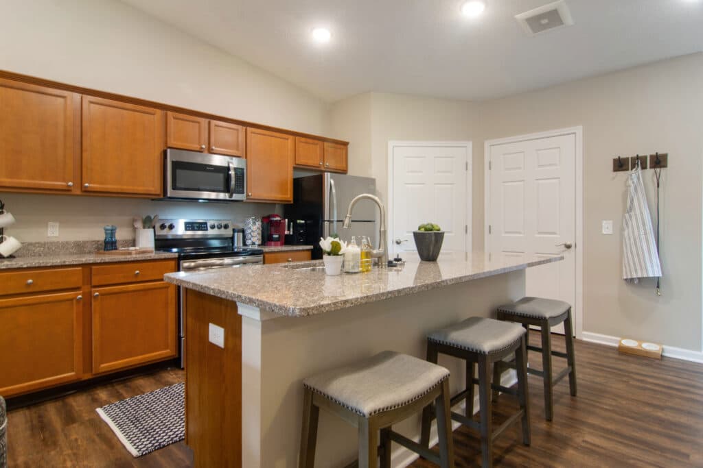 Modern kitchen with wooden cabinets, stainless steel appliances, a granite island with three stools, and neutral-colored walls and flooring. Hooks and aprons hang on the wall near double doors.