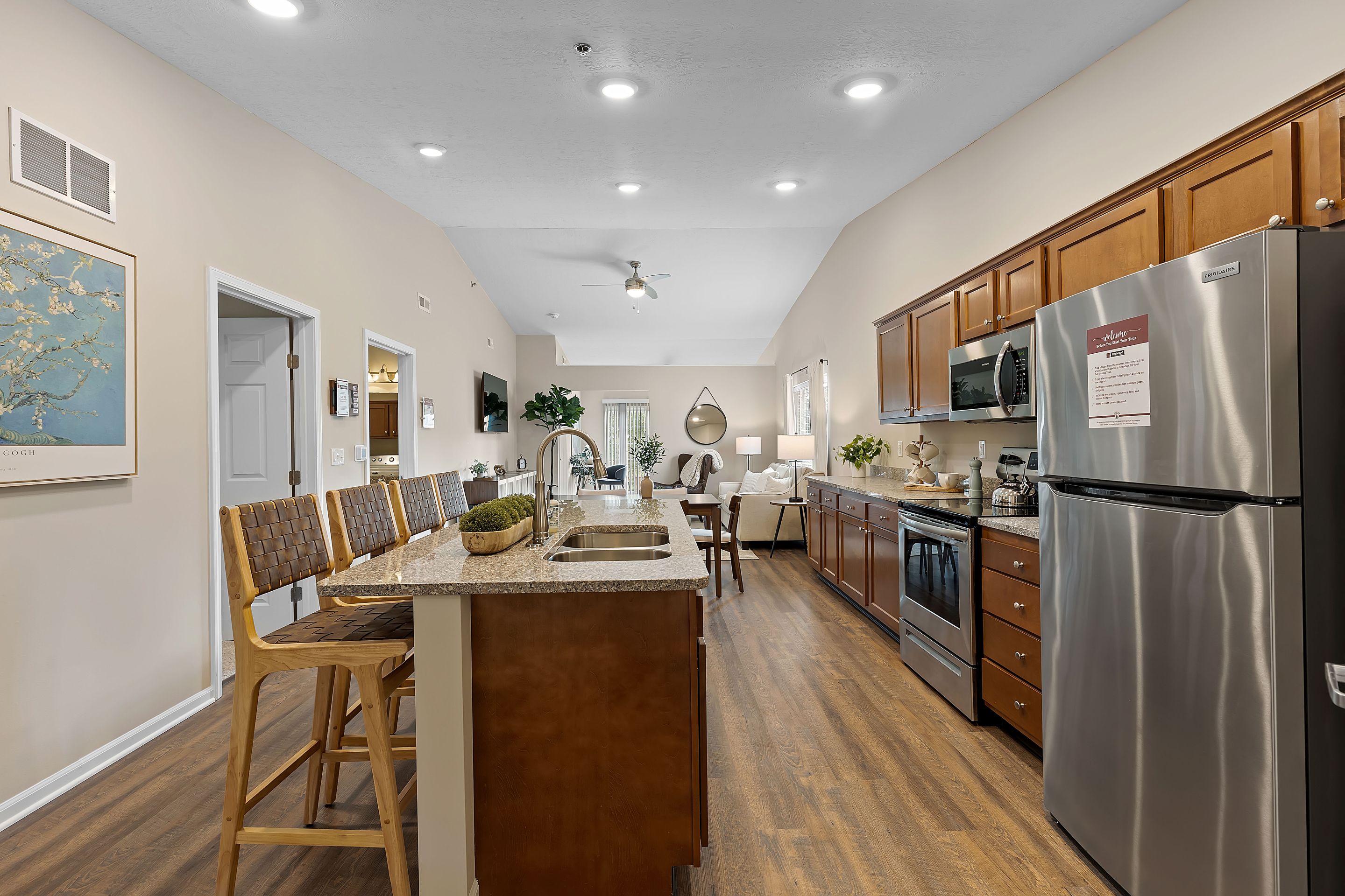 Open kitchen and living area with wood floors, stainless steel appliances, brown cabinets, bar seating, and neutral decor. Dining and living spaces visible in the background.