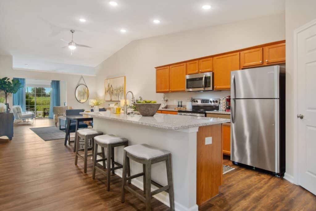 Modern kitchen with wooden cabinets, stainless steel appliances, a granite countertop island with three stools, and an open layout extending to a living area with large windows.
