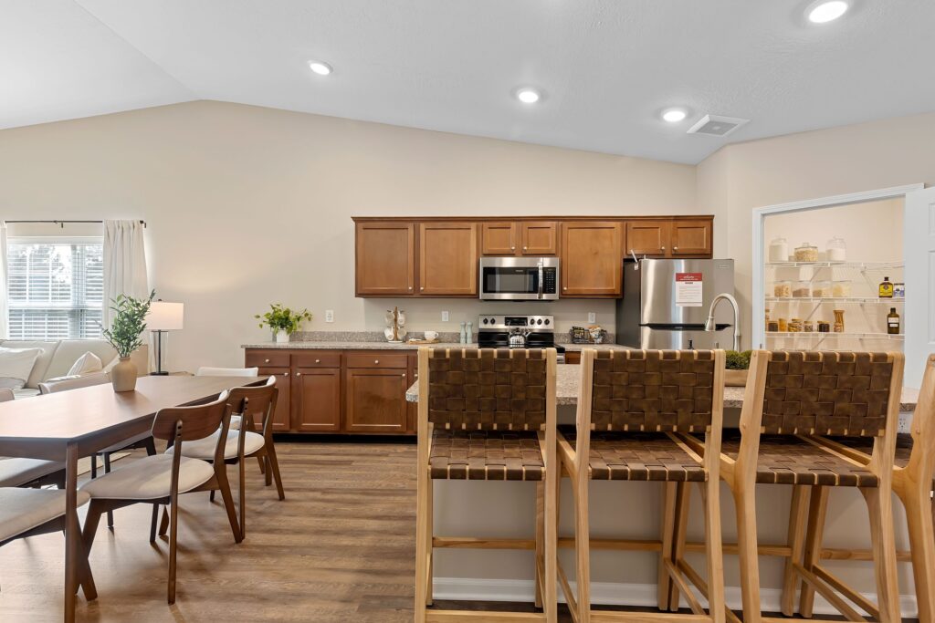 Modern kitchen and dining area with wooden cabinets, stainless steel appliances, a pantry, woven-back barstools, and a wooden dining table with chairs.