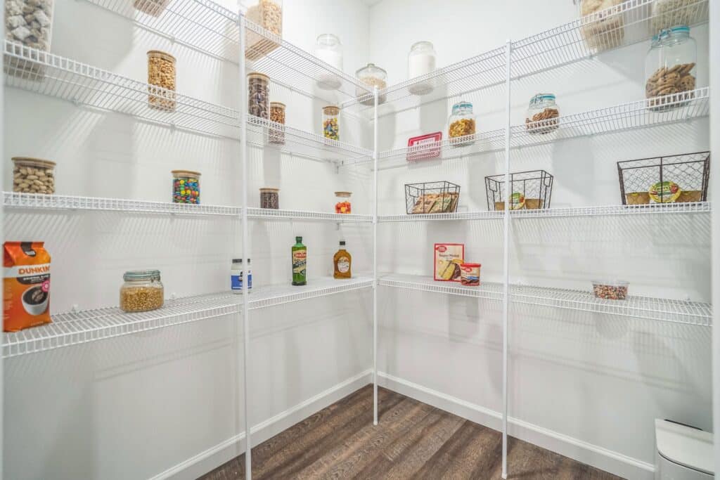 A pantry with white wire shelves holding assorted jars, baskets, and a few food items, organized against white walls with a wood floor.