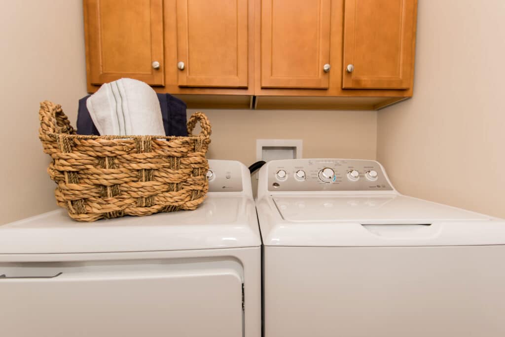 A laundry room with a washer and dryer, a woven basket with folded towels on top, and wooden cabinets mounted above.