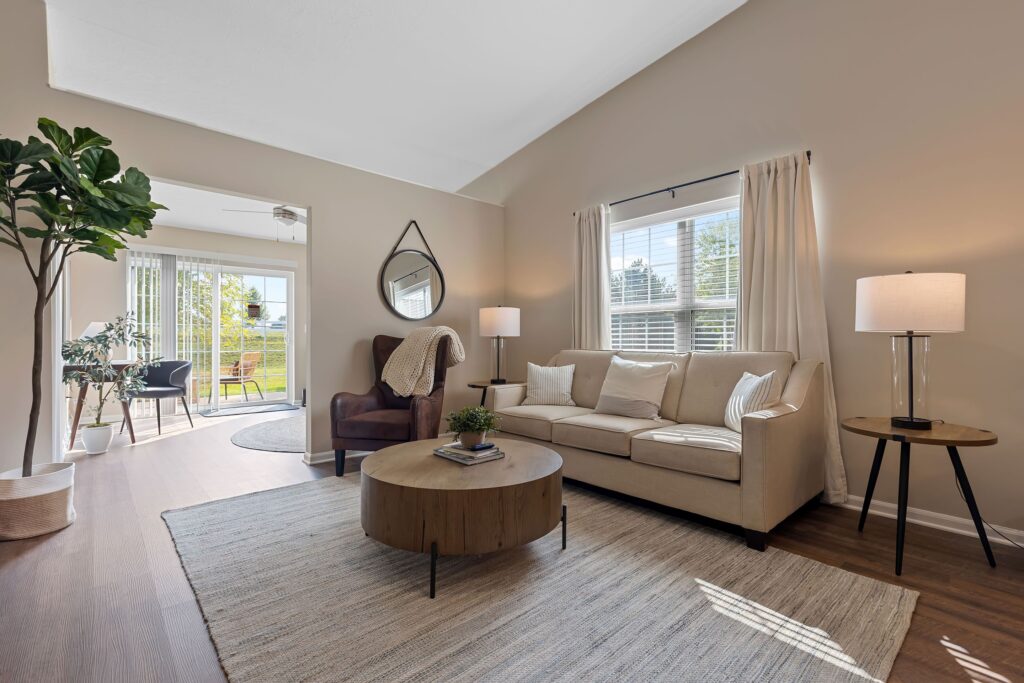 A modern living room with a beige sofa, round coffee table, armchair, and large window. A mirror hangs on the wall, and a sunroom with patio doors is visible in the background.