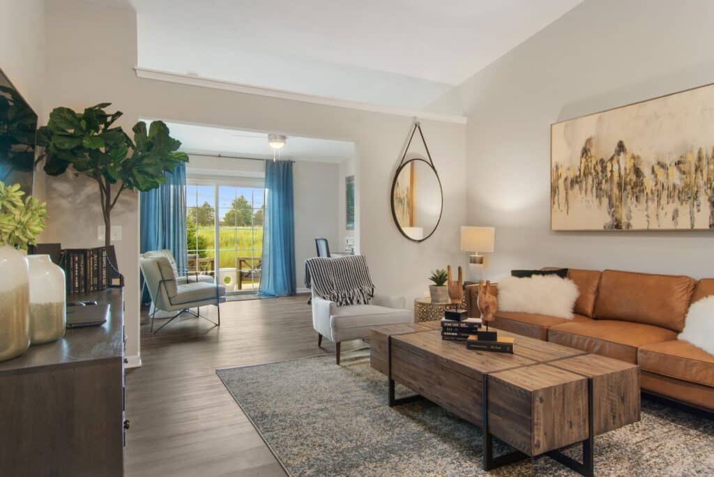 Modern living room with a brown sofa, wooden coffee table, neutral decor, large indoor plant, and a view into a sunroom with blue curtains and chairs.