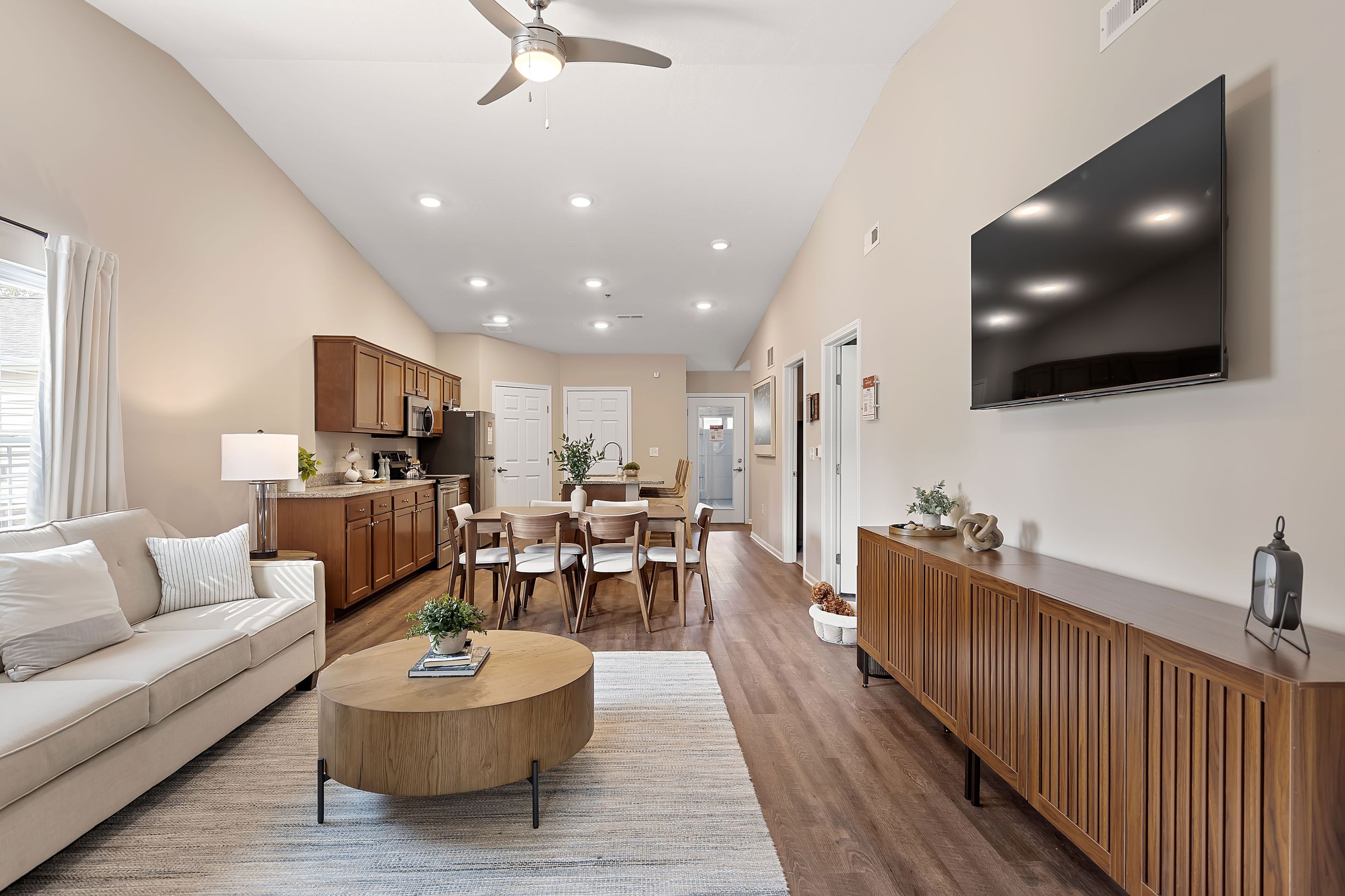 Open-concept living and dining area with a beige sofa, round coffee table, wooden dining set, wall-mounted TV, and wood cabinets in a bright, modern room.