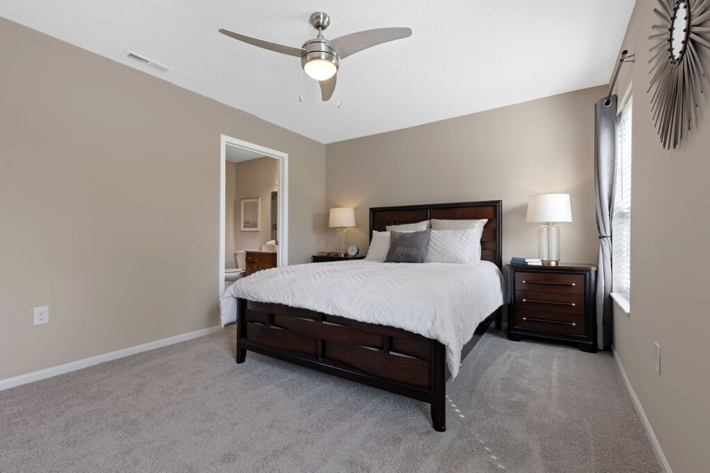 A modern bedroom with a dark wood bed, white bedding, matching nightstands with lamps, gray carpet, beige walls, and a ceiling fan with light.