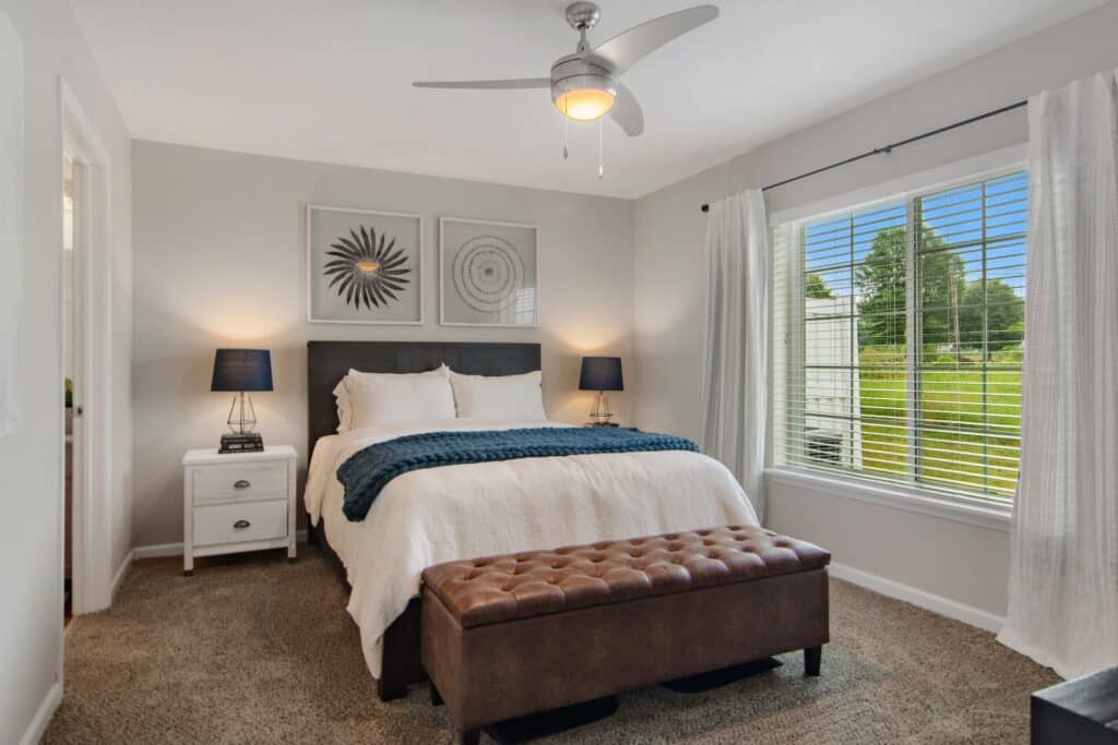A neatly made bed with white bedding, a dark headboard, and a brown bench in a bedroom with two nightstands, lamps, and a large window with white curtains.