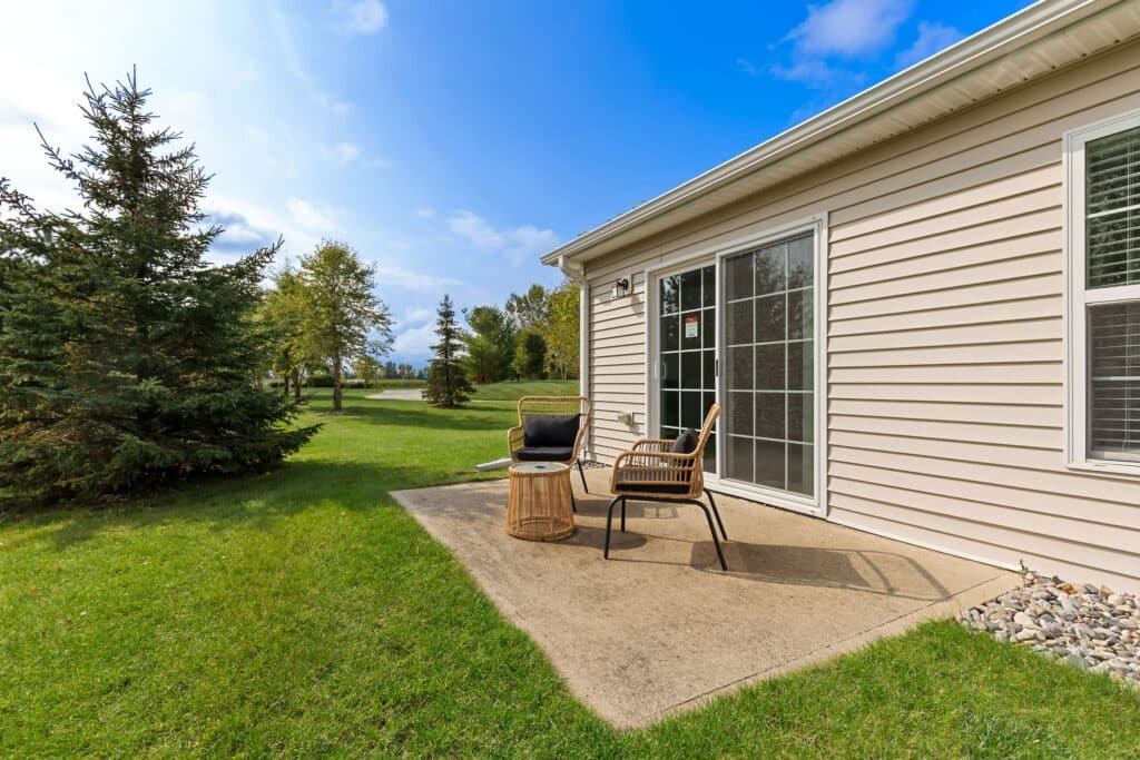 A small concrete patio with two wicker chairs and a barbecue grill beside a beige house, surrounded by grass and trees under a partly cloudy sky.
