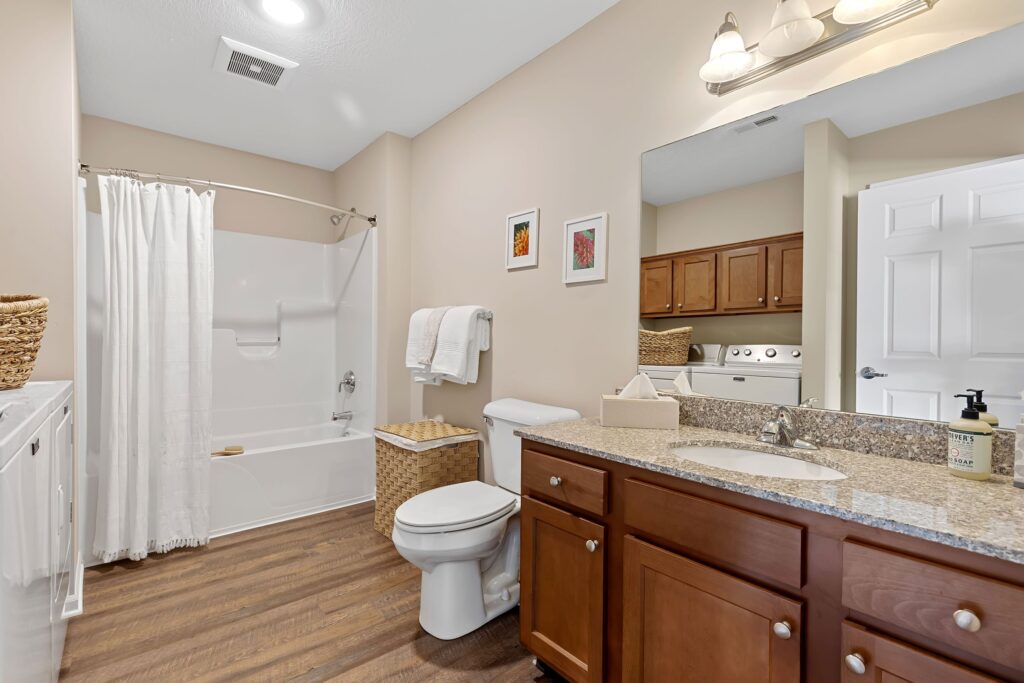 Modern bathroom with a granite countertop, wooden cabinets, toilet, bathtub with shower curtain, laundry area, and neutral wall decor.