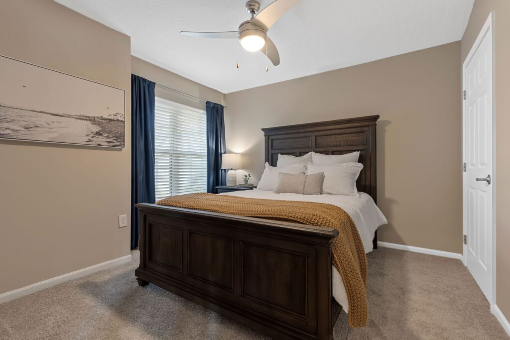 A bedroom with a dark wooden bed, white bedding, mustard blanket, nightstand with lamp, blue curtains, large window, ceiling fan, and beige walls.