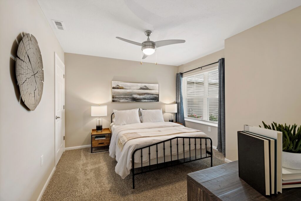 A neatly made bed with white linens is centered between two nightstands in a carpeted bedroom with beige walls, a ceiling fan, and a window with dark curtains.