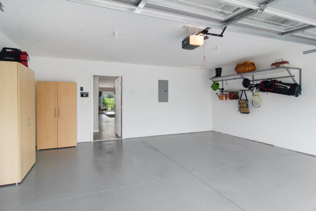 Clean, empty garage with gray floor, white walls, a wooden storage cabinet, and wall-mounted shelves holding various items. An open door leads to the interior of a house.