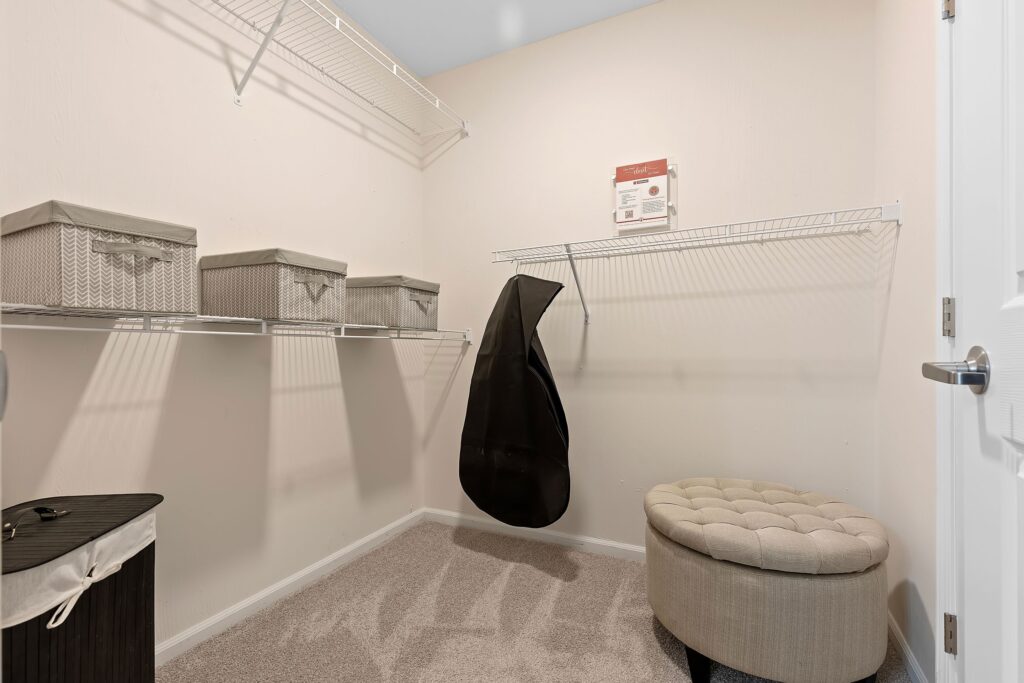 A walk-in closet with beige walls, wire shelves holding storage boxes, a hanging garment bag, a round tufted ottoman, and a laundry hamper on a carpeted floor.