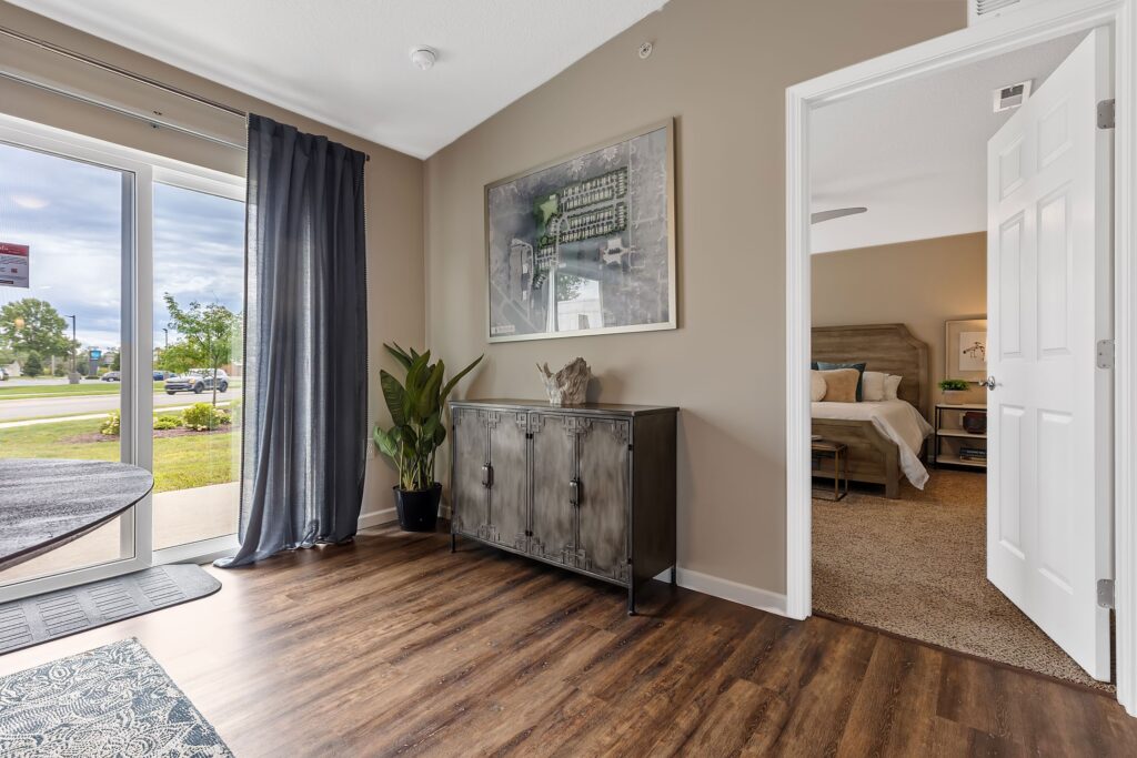 Modern apartment interior with wood flooring, a metal cabinet, large wall art, potted plant, and a view into a carpeted bedroom; large window brings in natural light.