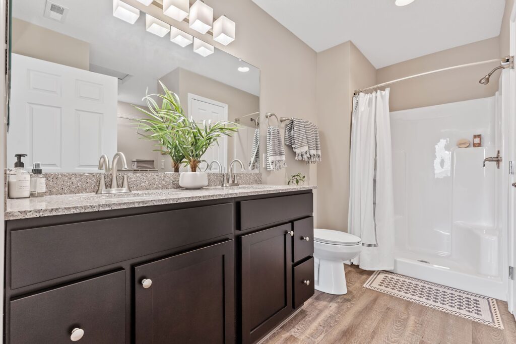 Modern bathroom with double sink vanity, dark cabinets, granite countertop, indoor plant, toilet, shower with curtain, towel racks, and wood-look flooring.