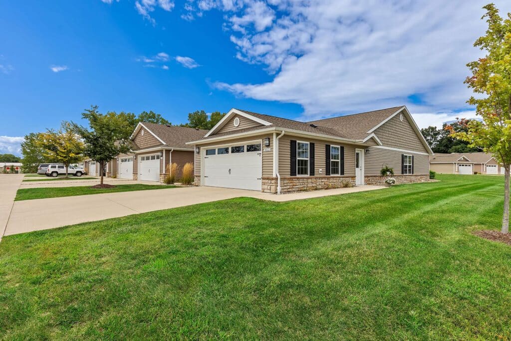 Single-story suburban house with beige siding, white trim, and a two-car garage, situated on a corner lot with green grass and a clear, partly cloudy sky.