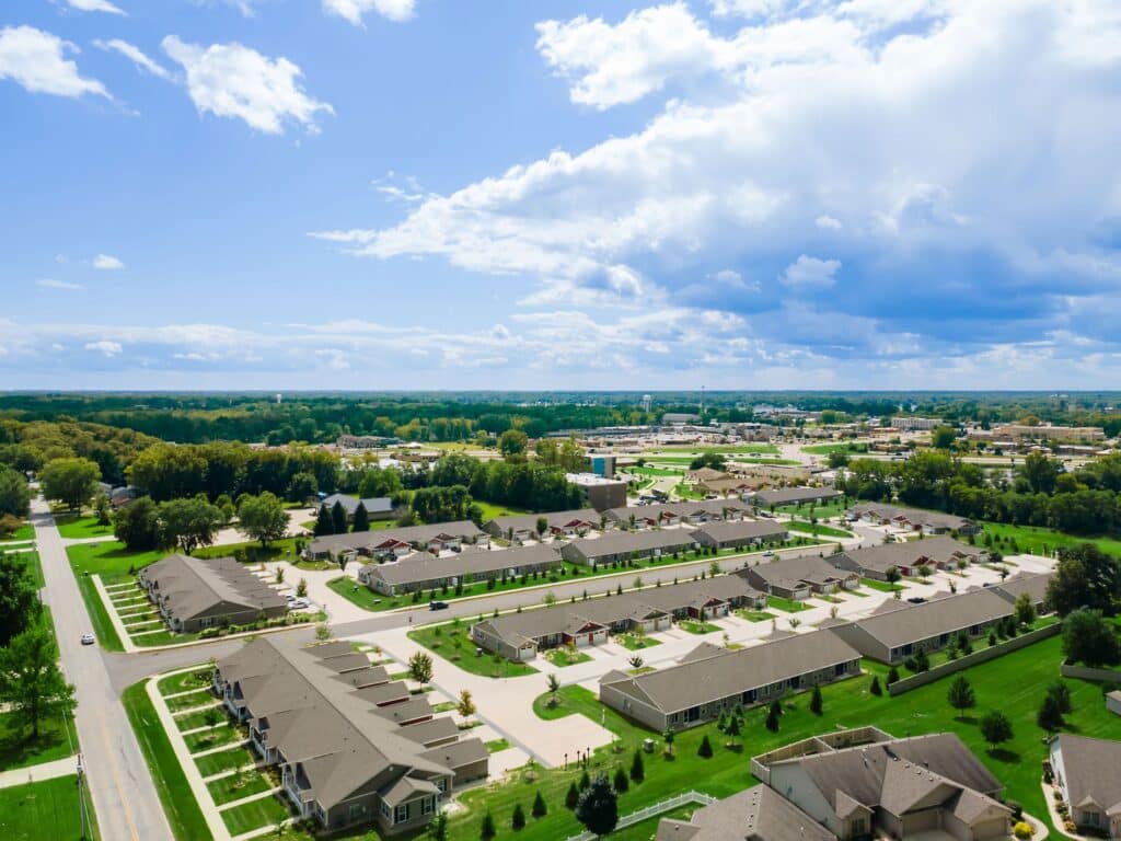 Aerial view of a residential neighborhood with rows of houses, green lawns, trees, roads, and partly cloudy sky.