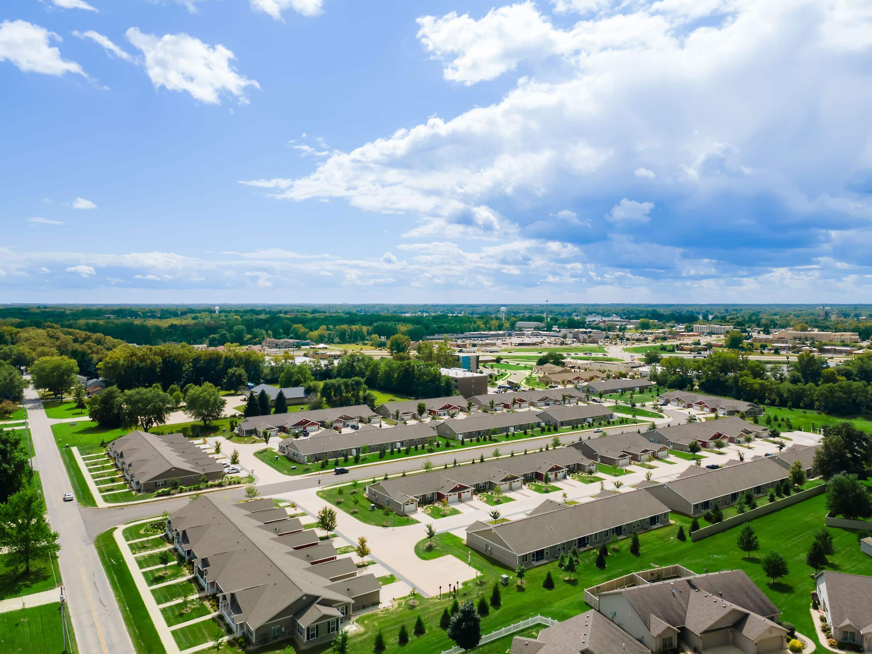 Aerial view of a residential neighborhood with rows of houses, green lawns, trees, roads, and partly cloudy sky.