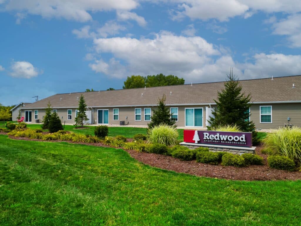 A single-story apartment building with a landscaped lawn and a sign reading "Redwood Apartment Neighborhoods" in front.