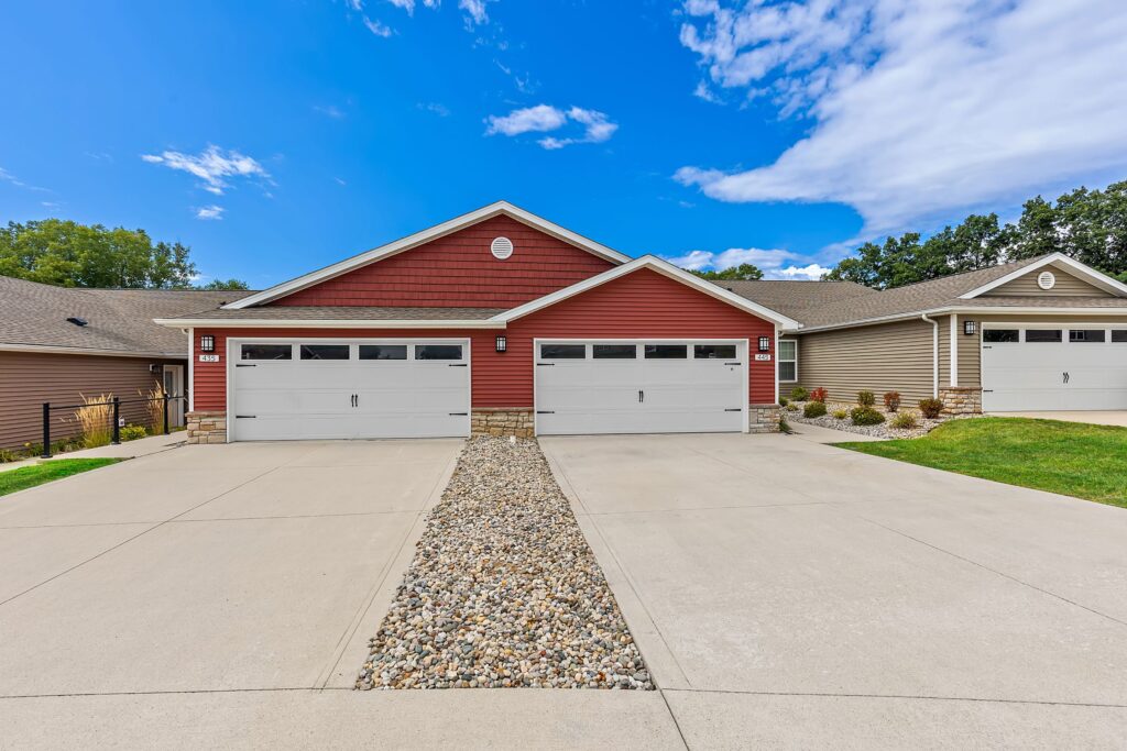 A duplex with two attached garages, red and tan siding, stone accents, and a concrete driveway with a central strip of decorative rocks under a partly cloudy sky.