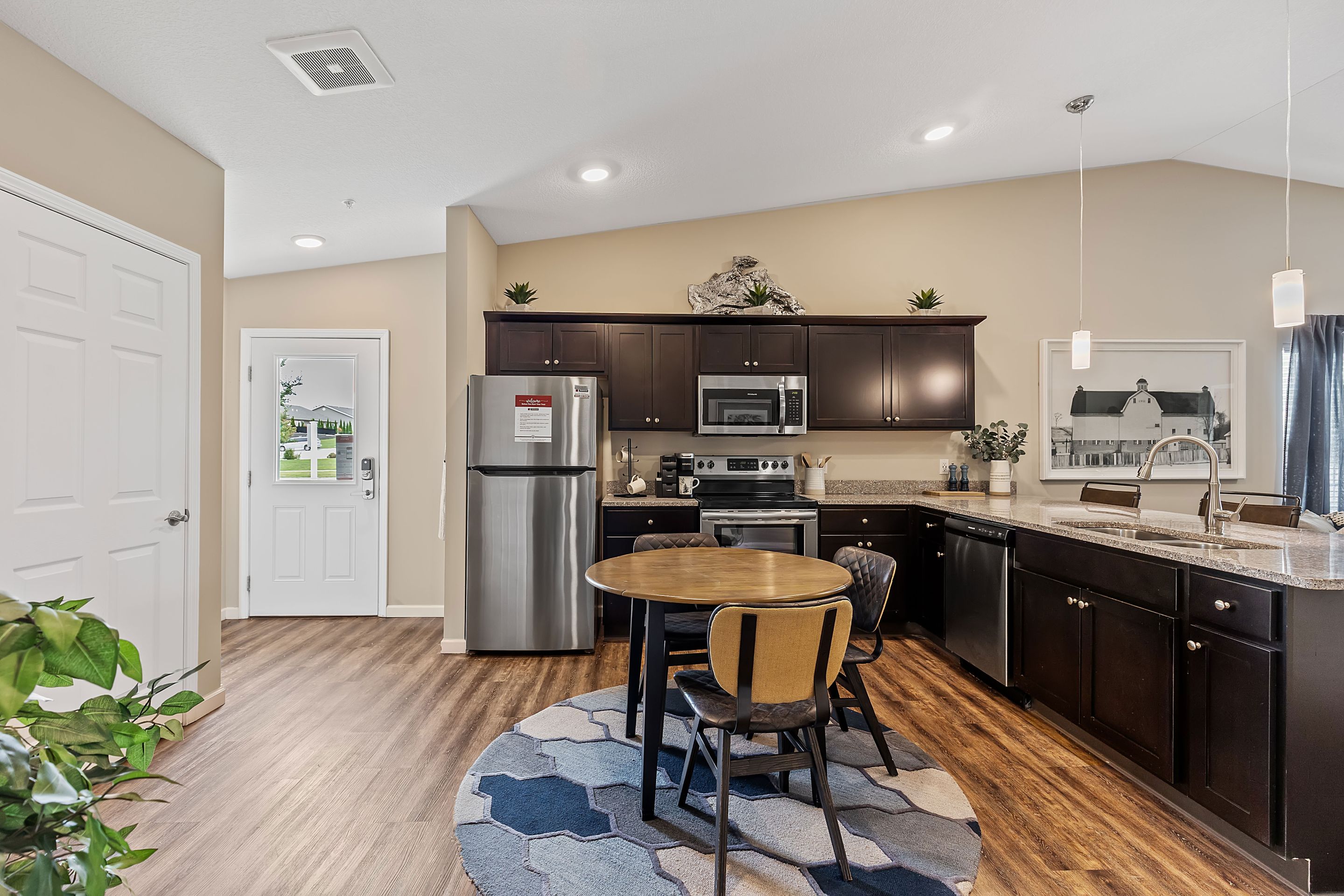 Modern kitchen with dark cabinets, stainless steel appliances, round dining table with chairs, and wood flooring. A door and window provide natural light.