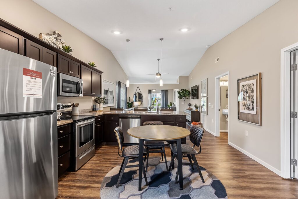 Modern kitchen with dark cabinets, stainless steel appliances, round dining table with four chairs on a patterned rug, and view into living area and hallway.