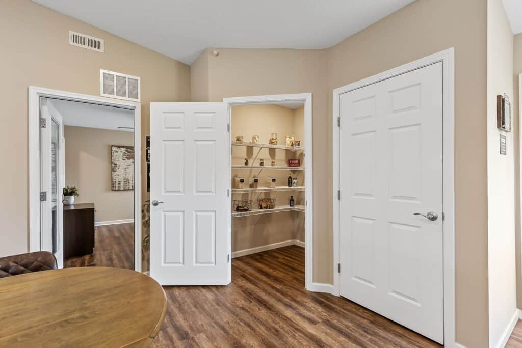 A small pantry with open shelves stocked with food items is seen between two white doors in a beige room with wood flooring.