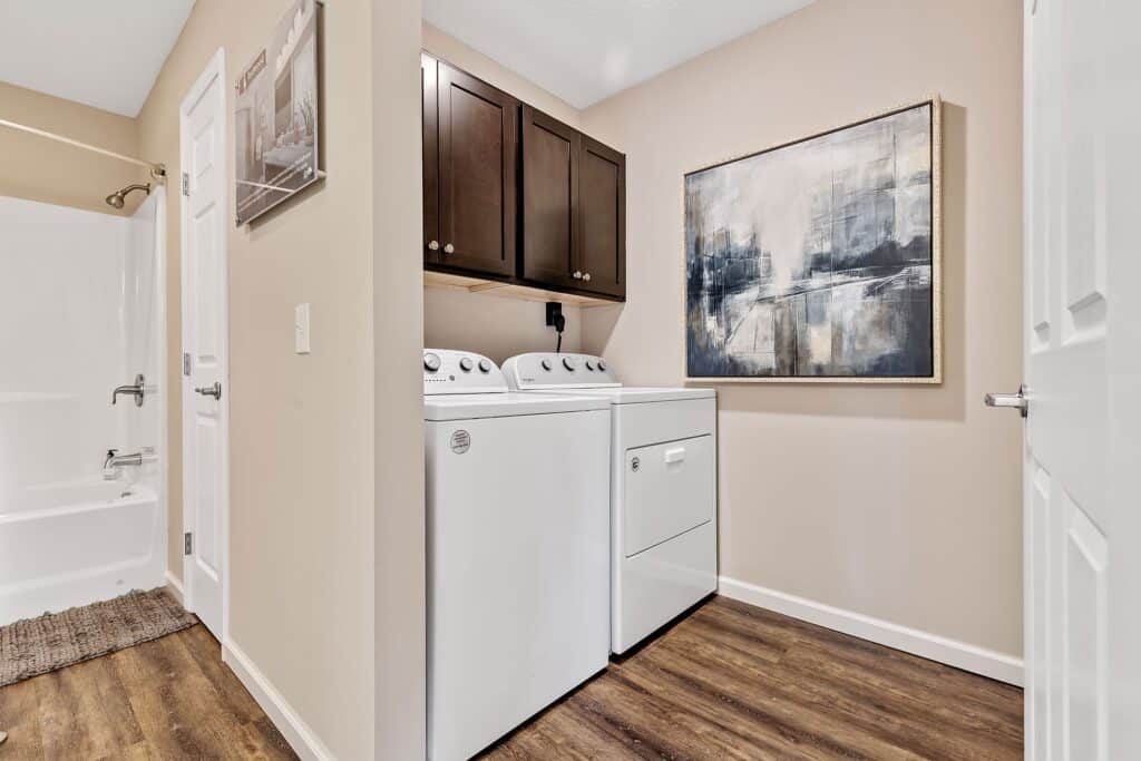 A laundry room with a washing machine and dryer, dark wood cabinets above, abstract wall art, and wood-look flooring next to a bathroom.