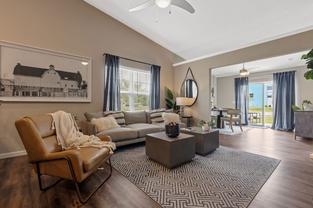 Modern living room with tan walls, neutral furniture, patterned rug, large windows with curtains, a plant, wall art, and an open dining area in the background.