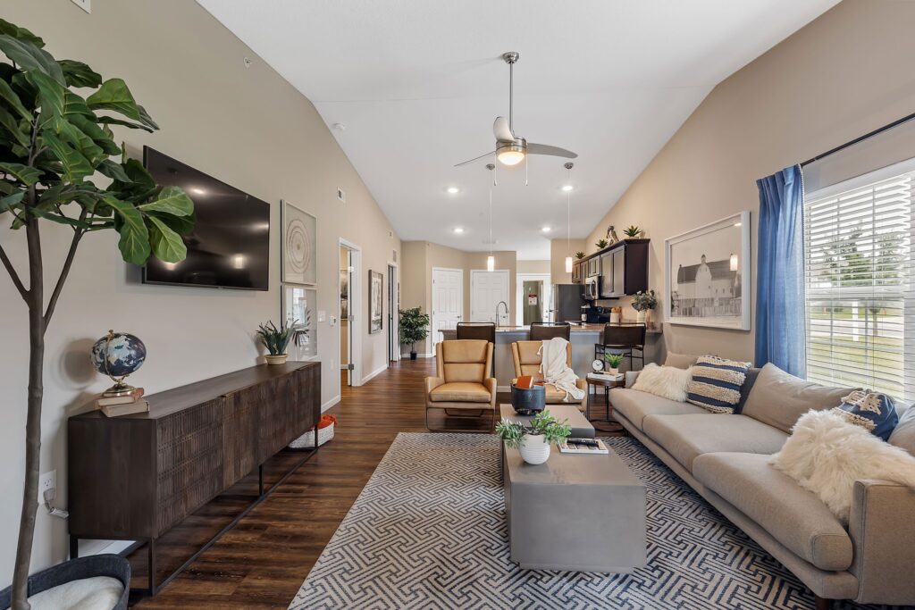 Modern living room with hardwood floors, a beige sofa, armchairs, a large TV, plants, and a patterned rug, connected to an open kitchen and dining area with high ceilings.