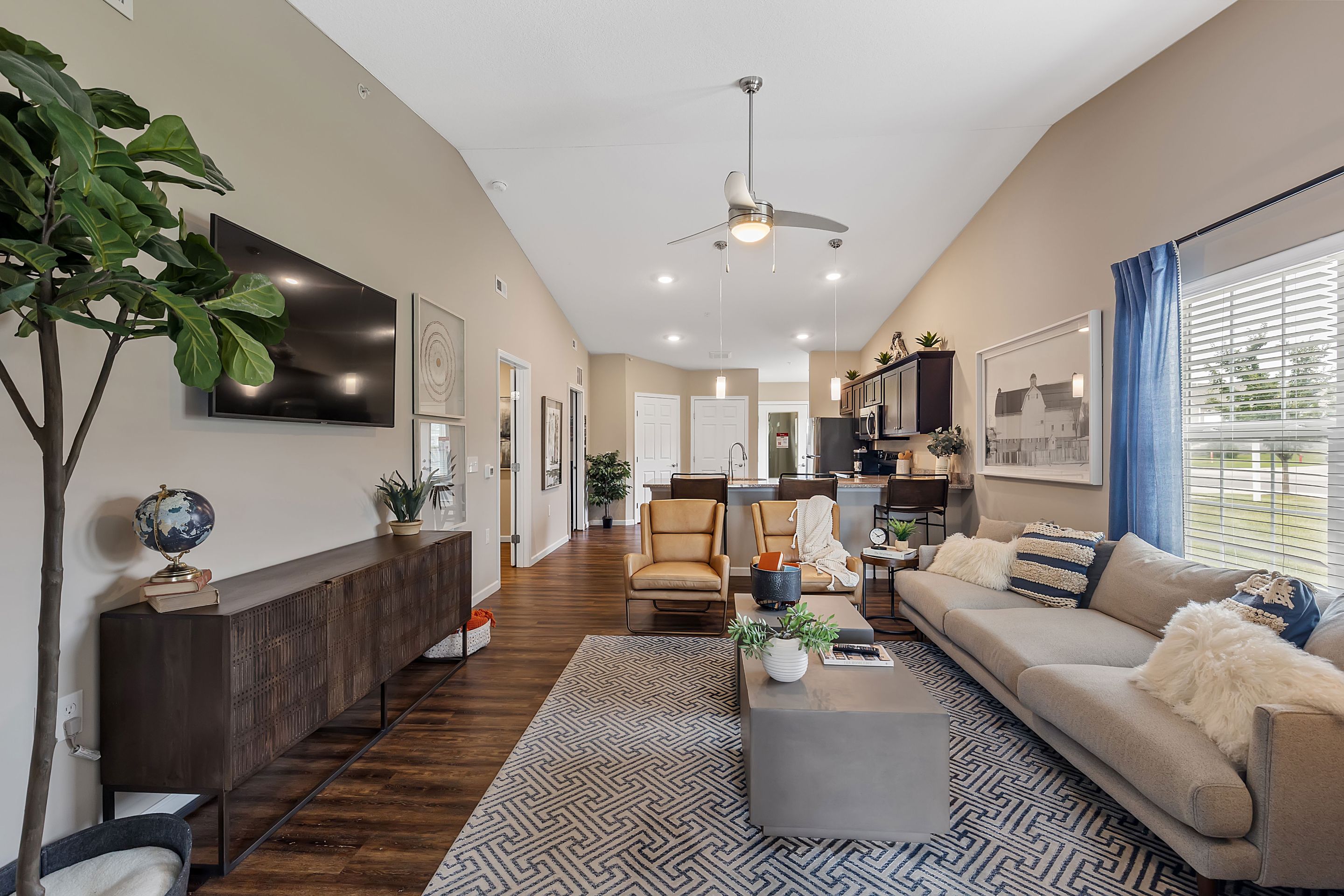 Modern living room with hardwood floors, a beige sofa, armchairs, a large TV, plants, and a patterned rug, connected to an open kitchen and dining area with high ceilings.