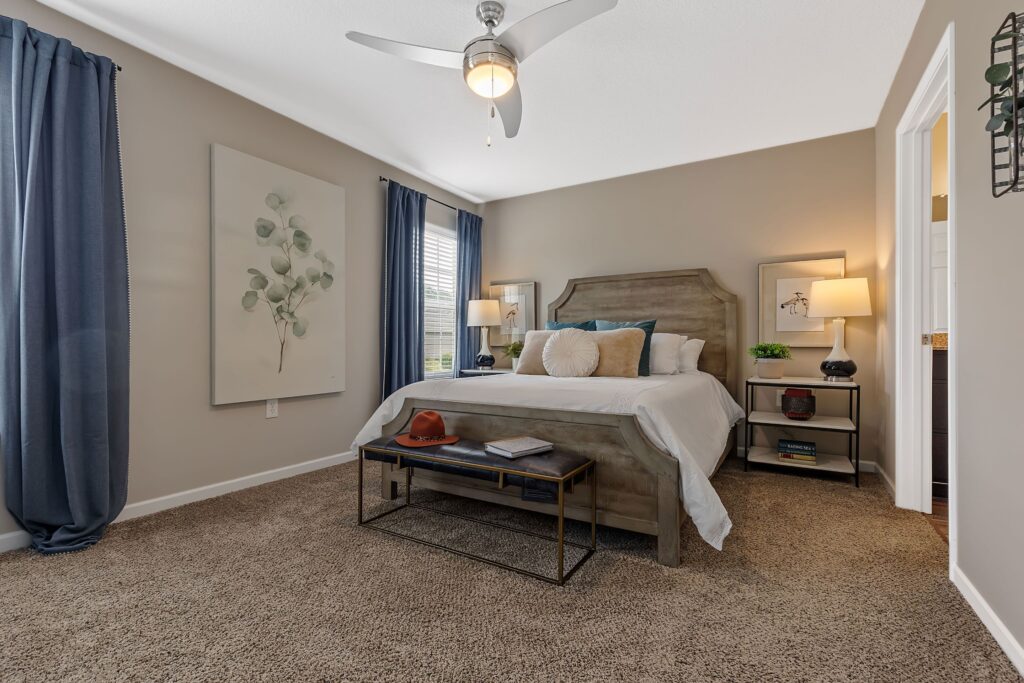 A neatly made bed with pillows sits against a beige wall in a bedroom with blue curtains, carpeted floor, a ceiling fan, and nightstands with lamps on each side.