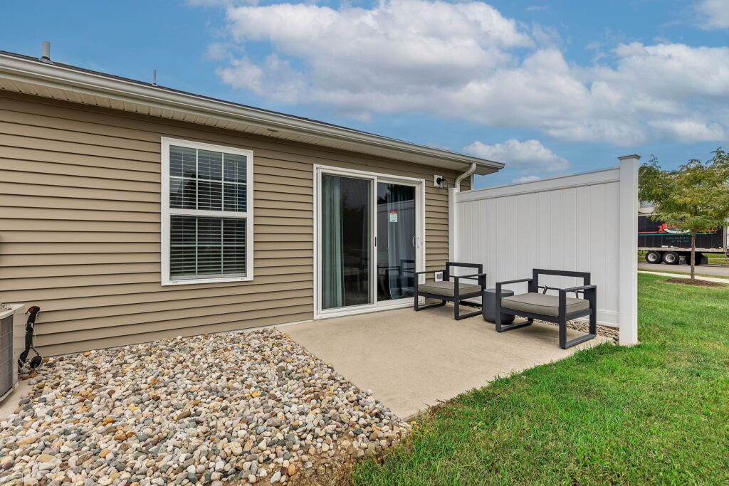 A small concrete patio with two black chairs, a white privacy fence, sliding glass door, beige siding, and a landscaped rock border next to a green lawn under a partly cloudy sky.