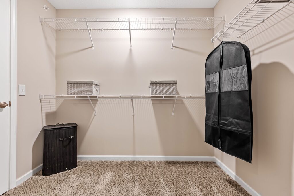 Walk-in closet with wire shelving, two storage boxes, a garment bag, and a black hamper on the carpeted floor.