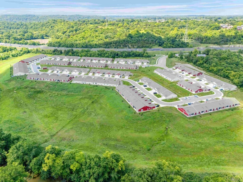 Aerial view of a residential neighborhood with rows of houses, surrounded by green fields, trees, and distant wooded hills under a partly cloudy sky.