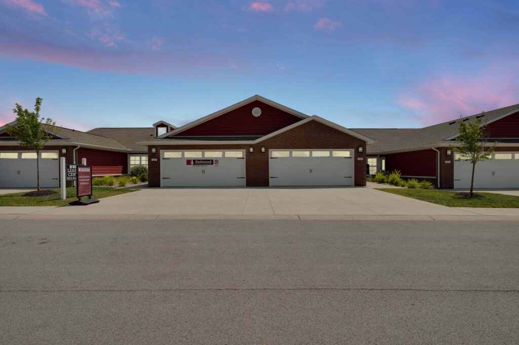 A row of attached red townhouse garages with white doors, brick accents, and a driveway, set against a blue sky with pink clouds.