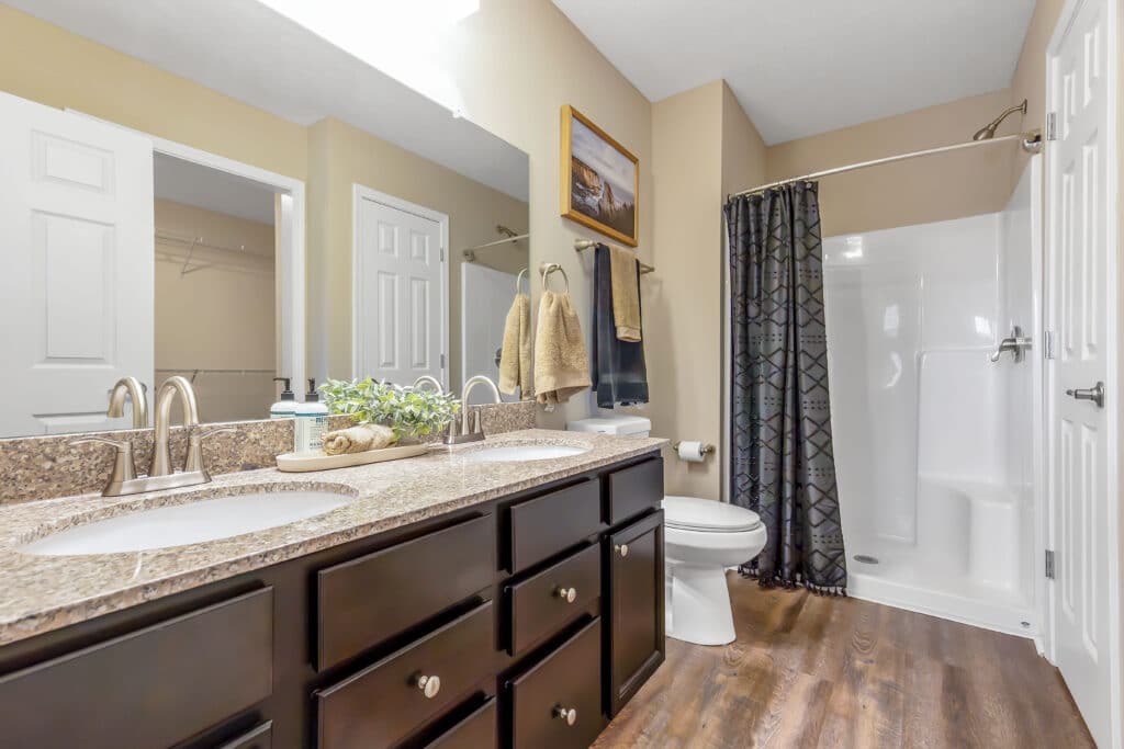 Modern bathroom with double sinks, granite countertop, dark cabinets, toilet, shower with patterned curtain, towels, and wall decor, all set on wood-look flooring.