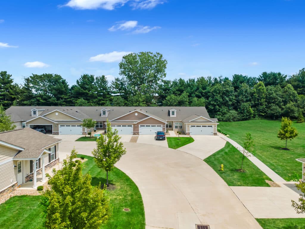 A row of suburban townhouses with driveways, garages, and well-maintained lawns, set along a curved street with trees and greenery in the background.