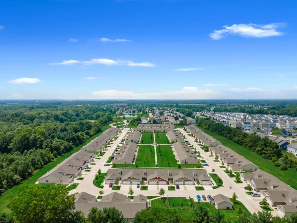 Aerial view of a suburban neighborhood with rows of houses, green lawns, and surrounding trees under a clear blue sky.